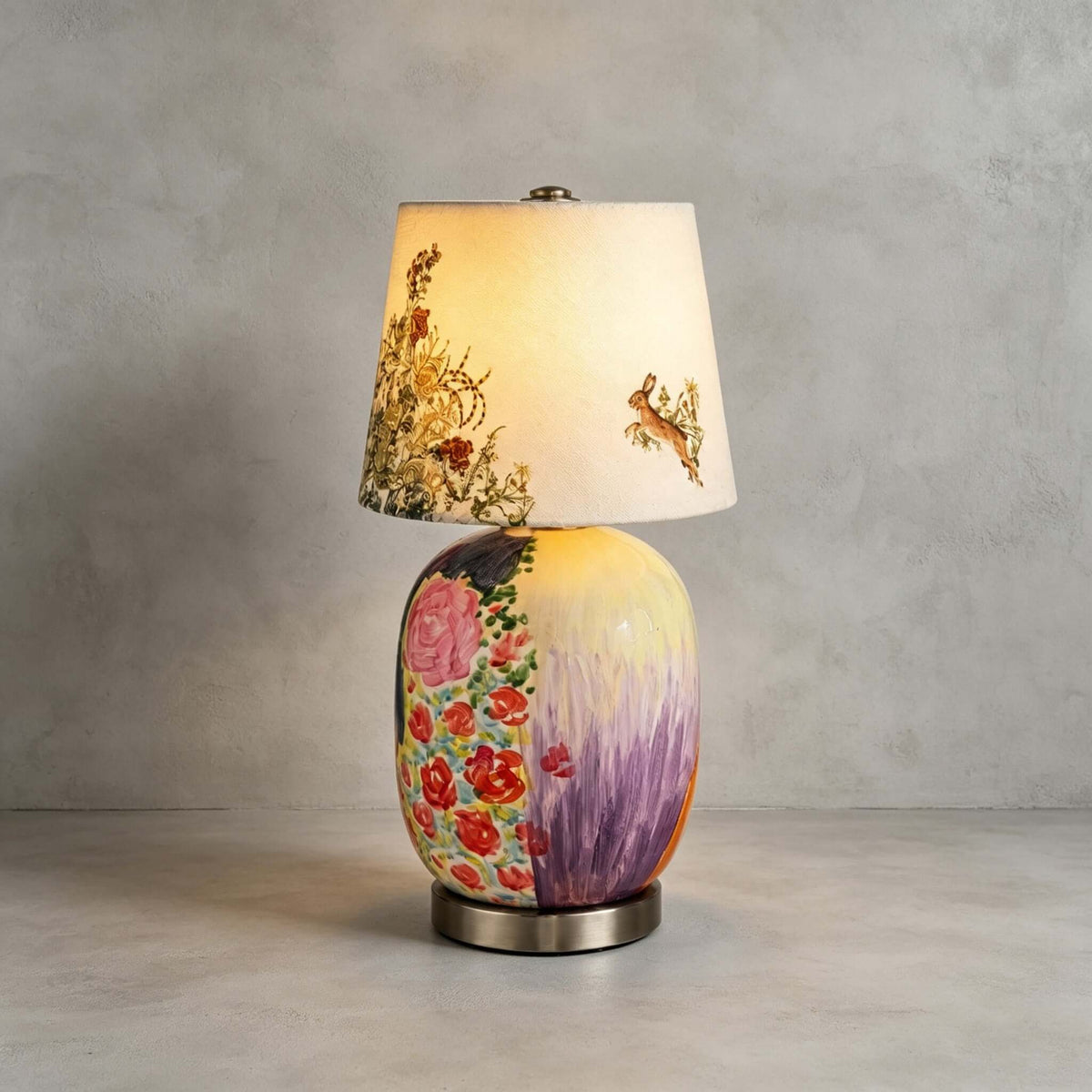 This Clérachic table lamp with a floral and lilac painted base stands on a concrete floor against a grey wall, its shade glowing.
