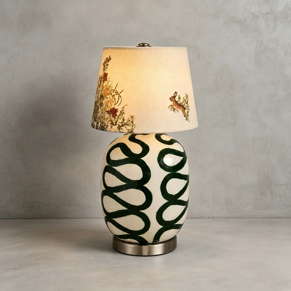 Green-looped Clérachic table lamp on a concrete floor against a grey wall, with the printed hare shade lit from within.