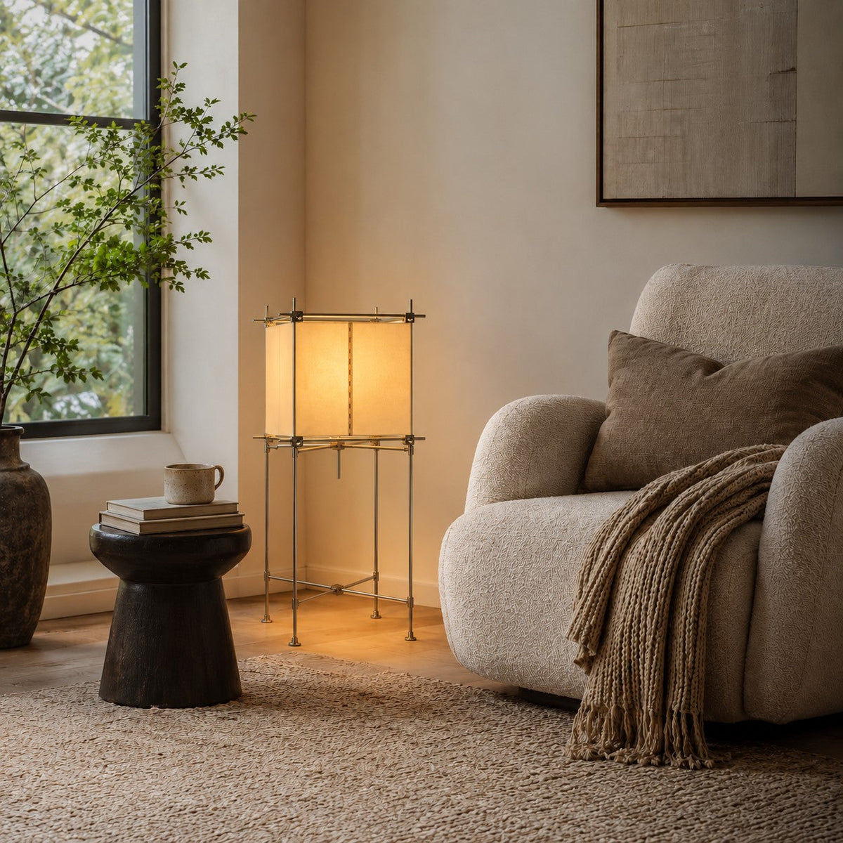 Lit Clérachic floor lamp with a paper cube shade beside a boucle armchair and a dark round side table in a window corner.