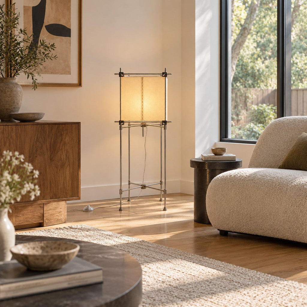A Clérachic floor lamp with a warm cube shade stands against a cream wall, above a wooden cabinet and beside a boucle chair.