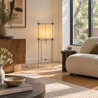 A Clérachic floor lamp with a warm cube shade stands against a cream wall, above a wooden cabinet and beside a boucle chair.