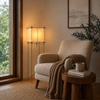 Warm light from a Clérachic cube floor lamp falls on a beige armchair with a knitted throw, beside a wood-framed window.