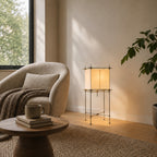 A Clérachic floor lamp with a paper shade lit in a bright corner, behind a round side table with a mug and two books.