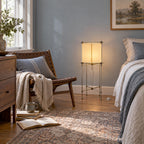 Clérachic cube floor lamp against a blue-grey wall in a bedroom, with a leather chair, a knitted throw and a bed to one side.