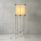 Full view of this Clérachic floor lamp, the cube parchment shade glowing on four metal legs with cross bracing, against a grey wall.