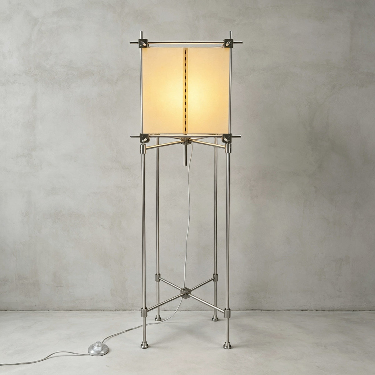 Full view of this Clérachic floor lamp, the cube parchment shade glowing on four metal legs with cross bracing, against a grey wall.