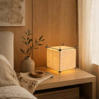 Next to a bed with linen bedding, this Clérachic parchment cube lamp lights an oak bedside table with a small vase and a woven throw.