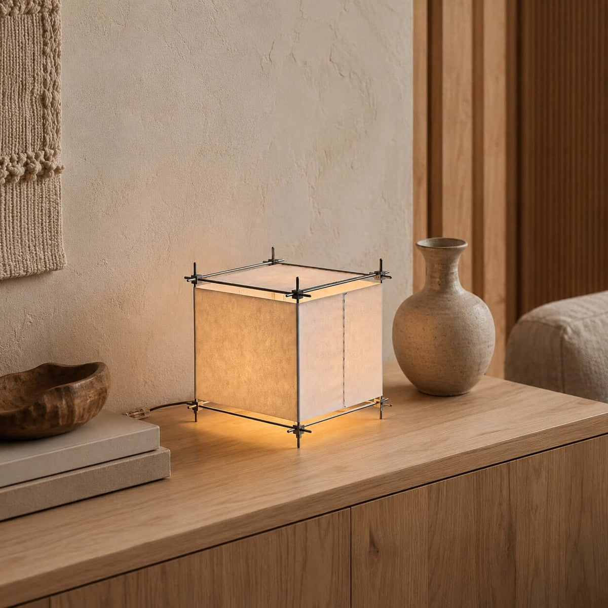 A Clérachic paper cube lamp stands on a pale sideboard with stacked books and a round-bellied vase, its warm light spilling beneath the frame.