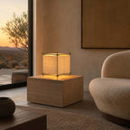 Placed on a travertine block beside a boucle armchair, this Clérachic paper cube lamp glows against a wall at sunset.