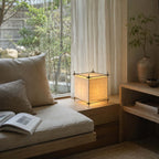 On a window seat with a cushion and open book, this Clérachic cube lamp glows beside sheer curtains and a garden view.