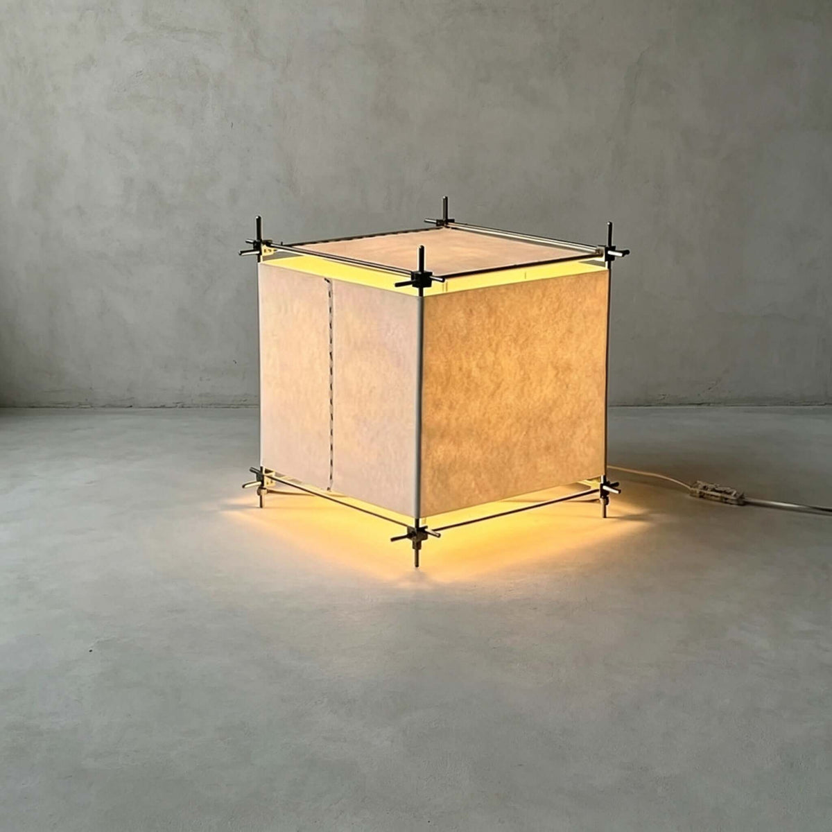 Standing alone on a grey concrete floor, this Clérachic cube lamp shows its parchment panels glowing, with the light spilling under the frame.