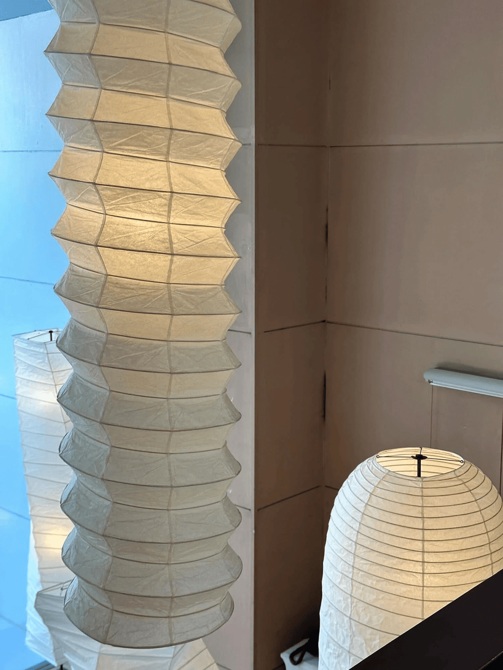 Clérachic pleated paper pendant light shown in close crop against a tan panelled wall, with a separate ribbed paper lamp standing on its base behind.