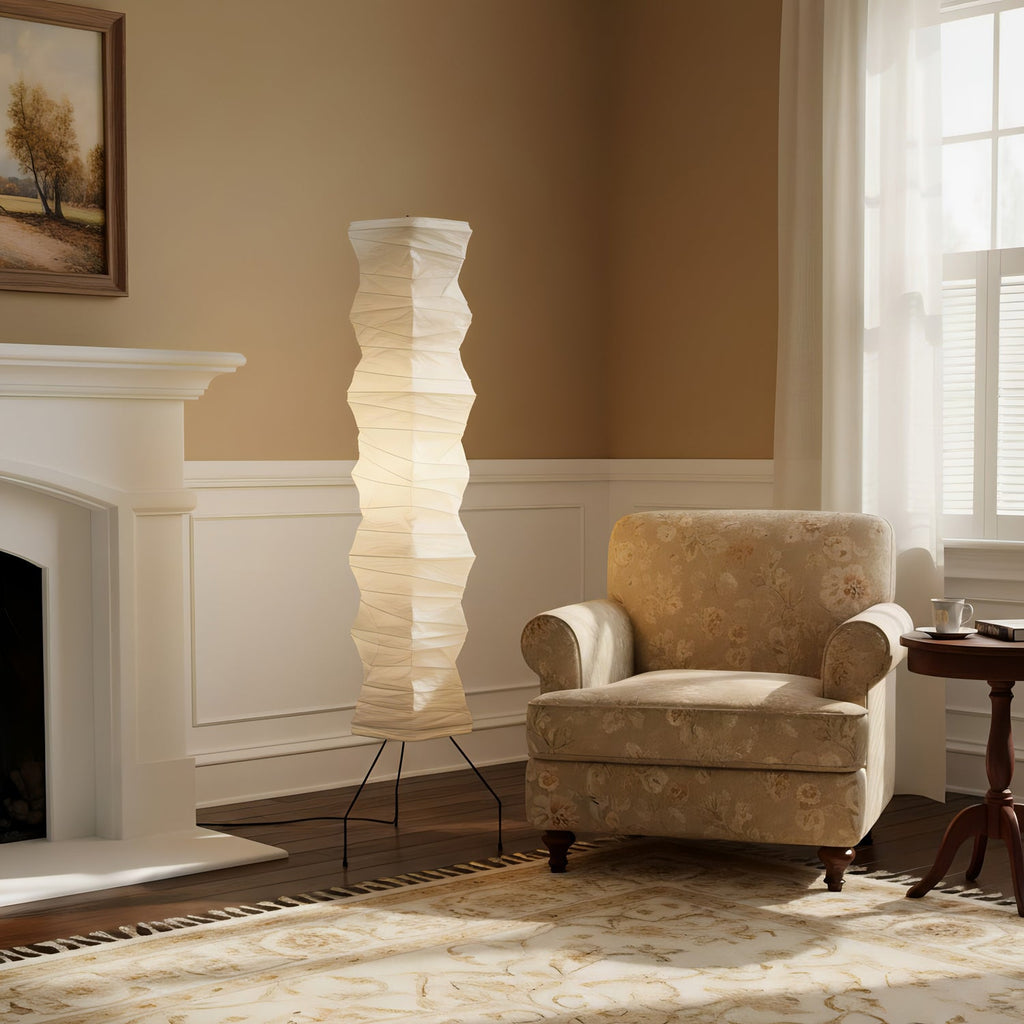 A Clérachic paper floor lamp glowing beside a white fireplace, with a floral armchair and a patterned rug in a cream room.