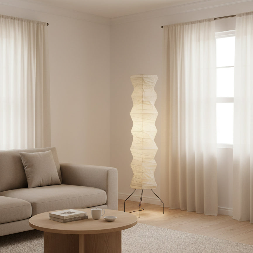 This Clérachic folded paper floor lamp stands beside a grey sofa and a round wooden table, with sheer curtains behind.