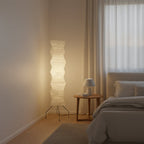 A Clérachic paper floor lamp lit beside a bed with grey bedding and a small round bedside table, with a window behind.