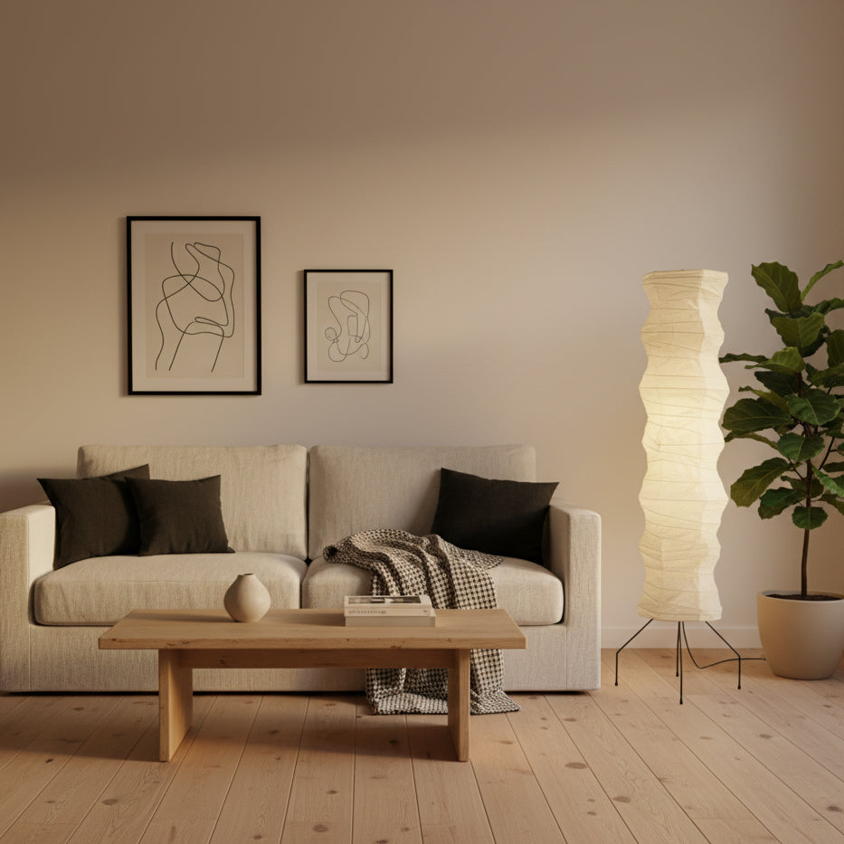 Clérachic tall paper floor lamp in a living room, beside a cream sofa with dark cushions and a leafy plant.