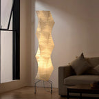 Close view of the lit, creased paper shade on this Clérachic floor lamp, in a living room with a window and a sofa.