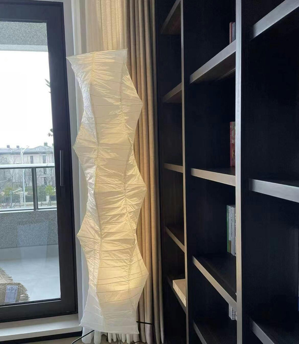 Tall Clérachic paper floor lamp glowing beside a glass door and a dark bookshelf, its shade folded in soft ridges.