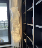 Tall Clérachic paper floor lamp glowing beside a glass door and a dark bookshelf, its shade folded in soft ridges.
