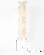 Clérachic paper floor lamp on a pale backdrop, its creased shade standing on three black legs with a cord and foot switch on the floor.