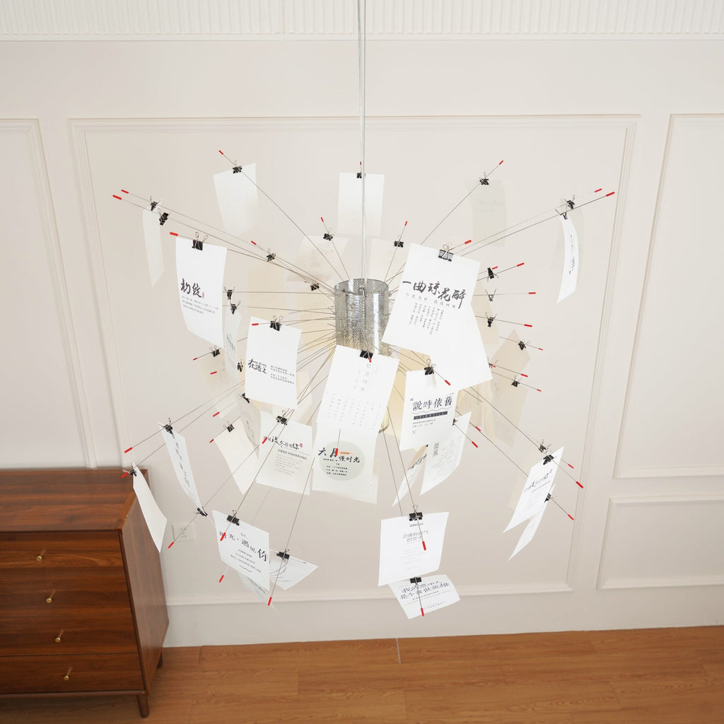 A Clérachic paper pendant with red-tipped wire arms and clipped white cards, hung low in a panelled room beside a wooden chest.