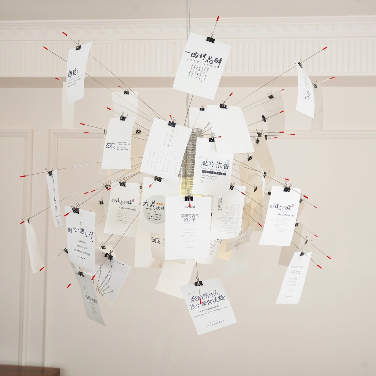 Close view of a Clérachic paper pendant with printed text cards clipped to red-tipped wire arms against a panelled wall.