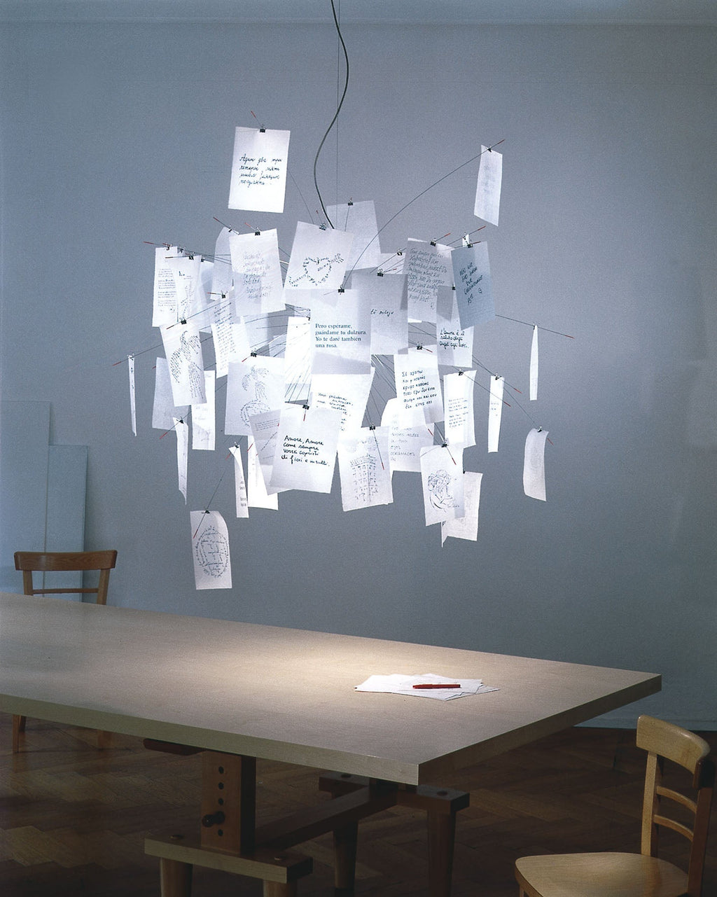 A Clérachic paper pendant above a long table in a grey-blue room, with handwritten notes clipped all over its frame.