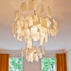 Warm light spills through layered paper notes on this Clérachic pendant, hung from a white ceiling above a pair of curtained windows.