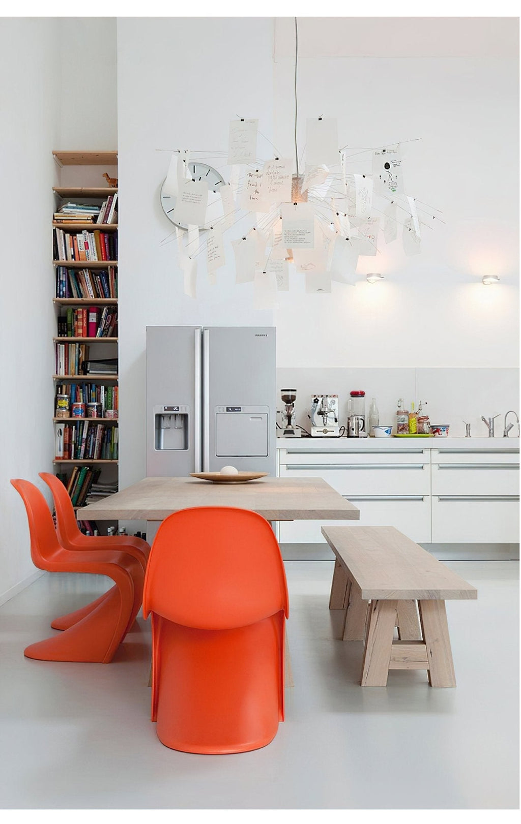 Above a pale wood table in a white kitchen with orange chairs, a Clérachic paper pendant glows high in the room.