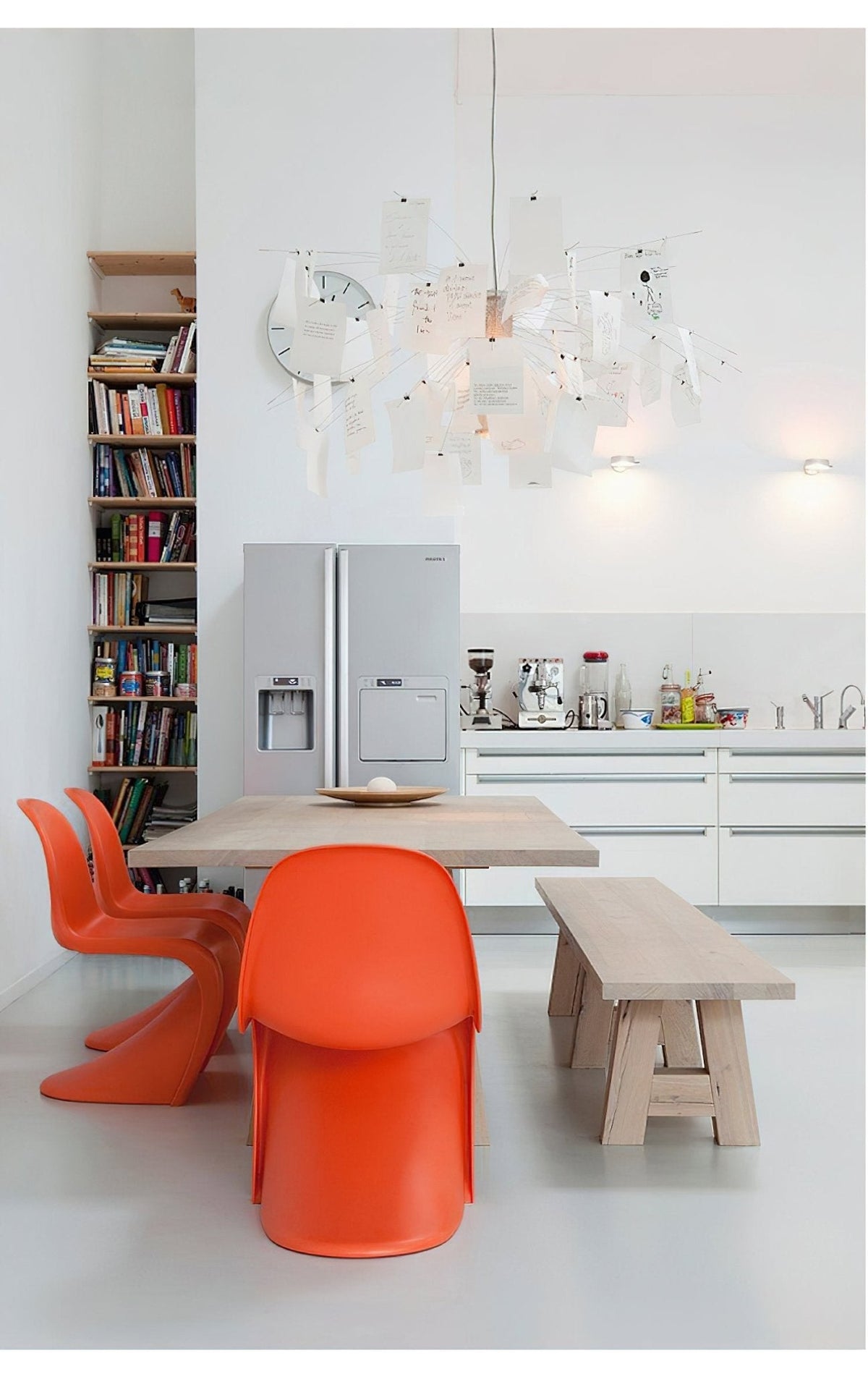 Above a pale wood table in a white kitchen with orange chairs, a Clérachic paper pendant glows high in the room.