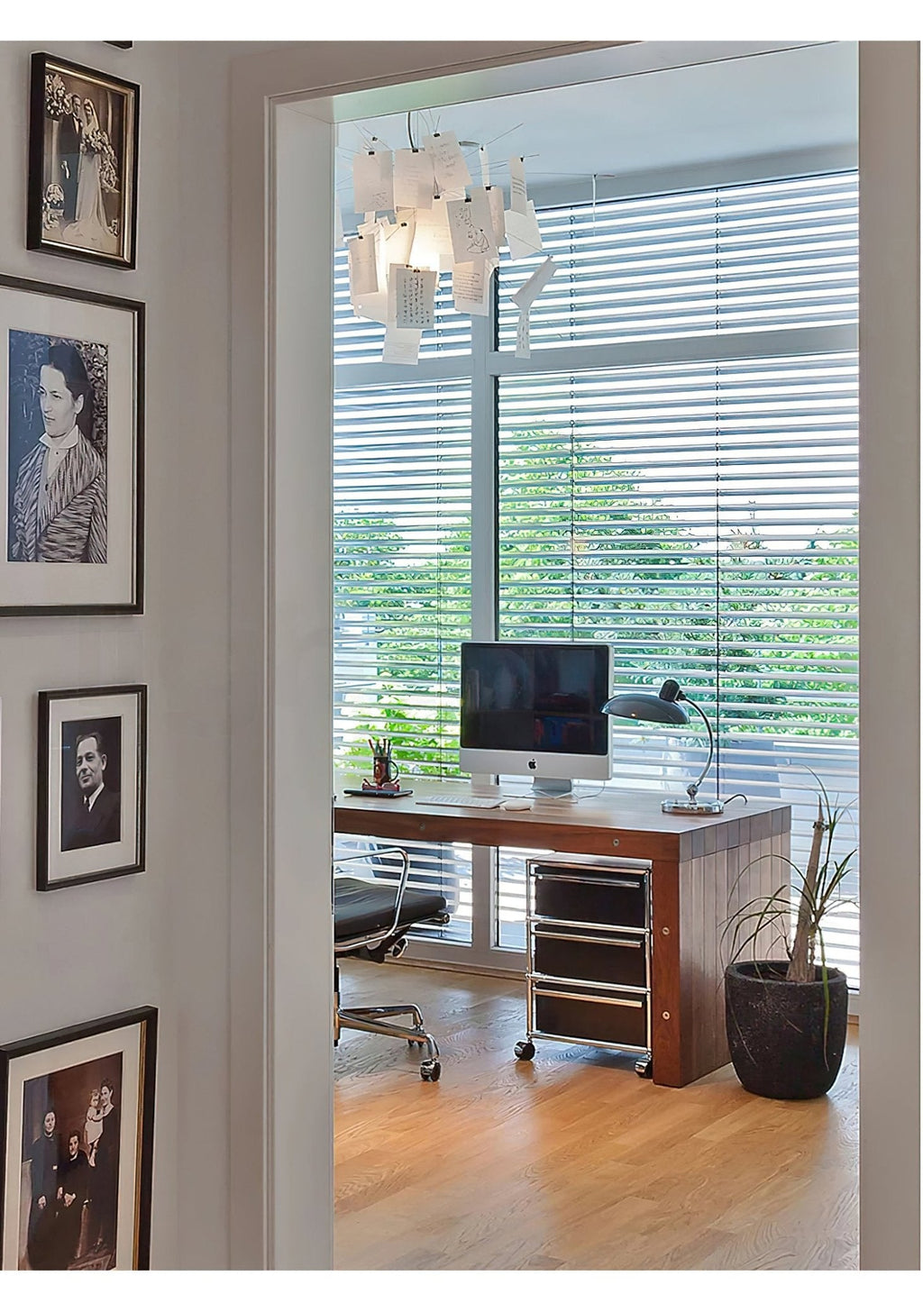 Through a doorway, a Clérachic paper pendant hangs in a study with framed portraits and a desk by a window.