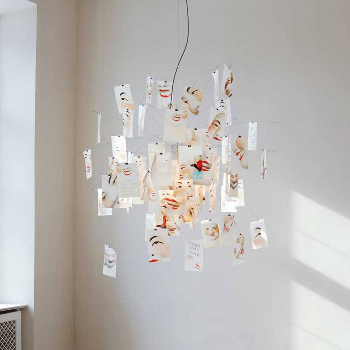 Cards with painted faces and red lips cover this Clérachic paper pendant, lit from within in a white corner with soft sunlight.