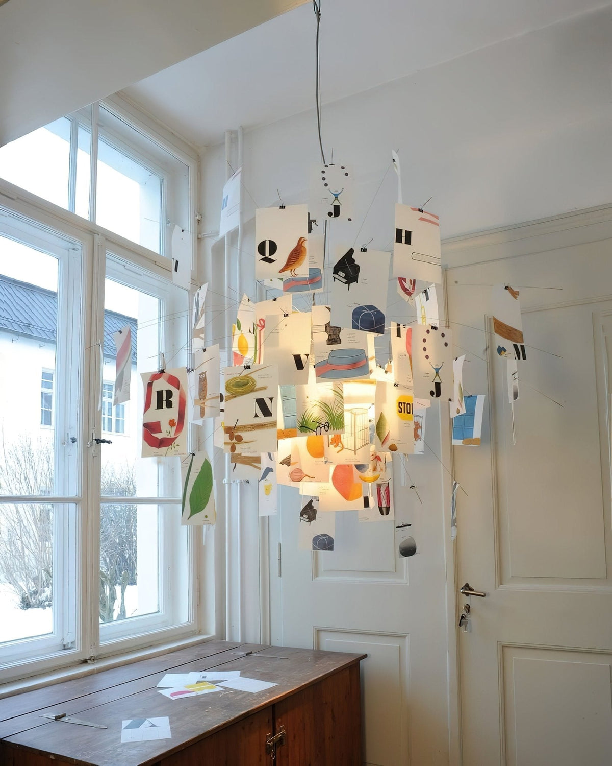 Lit warmly beside tall windows, a Clérachic pendant with printed letter cards and wire arms hangs over a wooden chest.