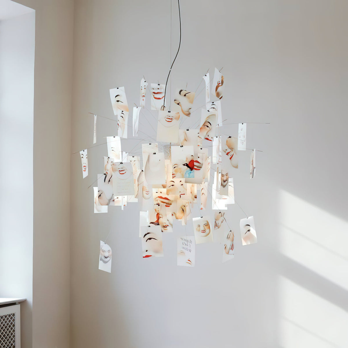 Cards printed with painted faces hang from this Clérachic paper pendant in a pale room with sunlight on the wall.