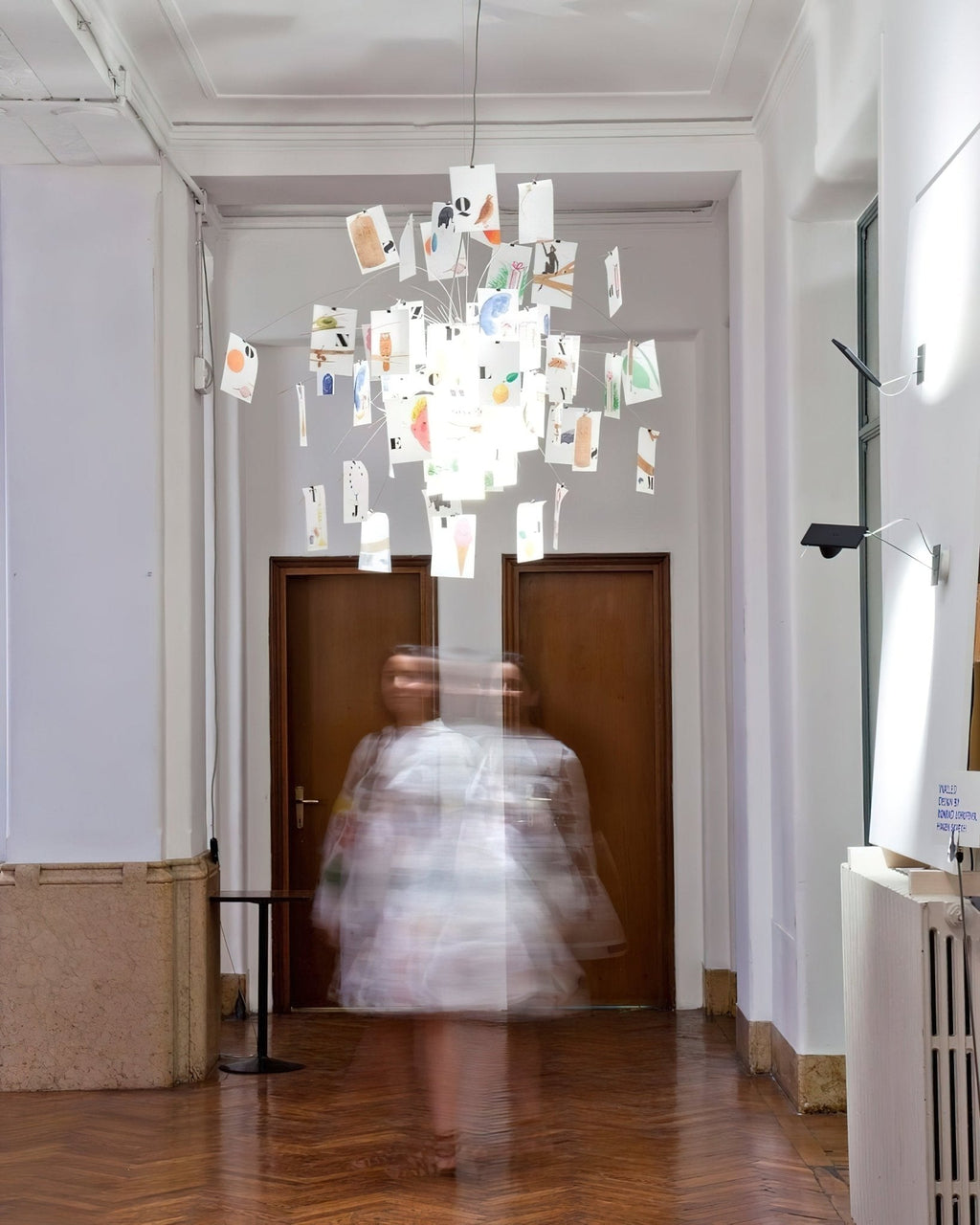 At the end of a long hall, a Clérachic paper pendant with colourful printed cards hangs above a blurred figure.