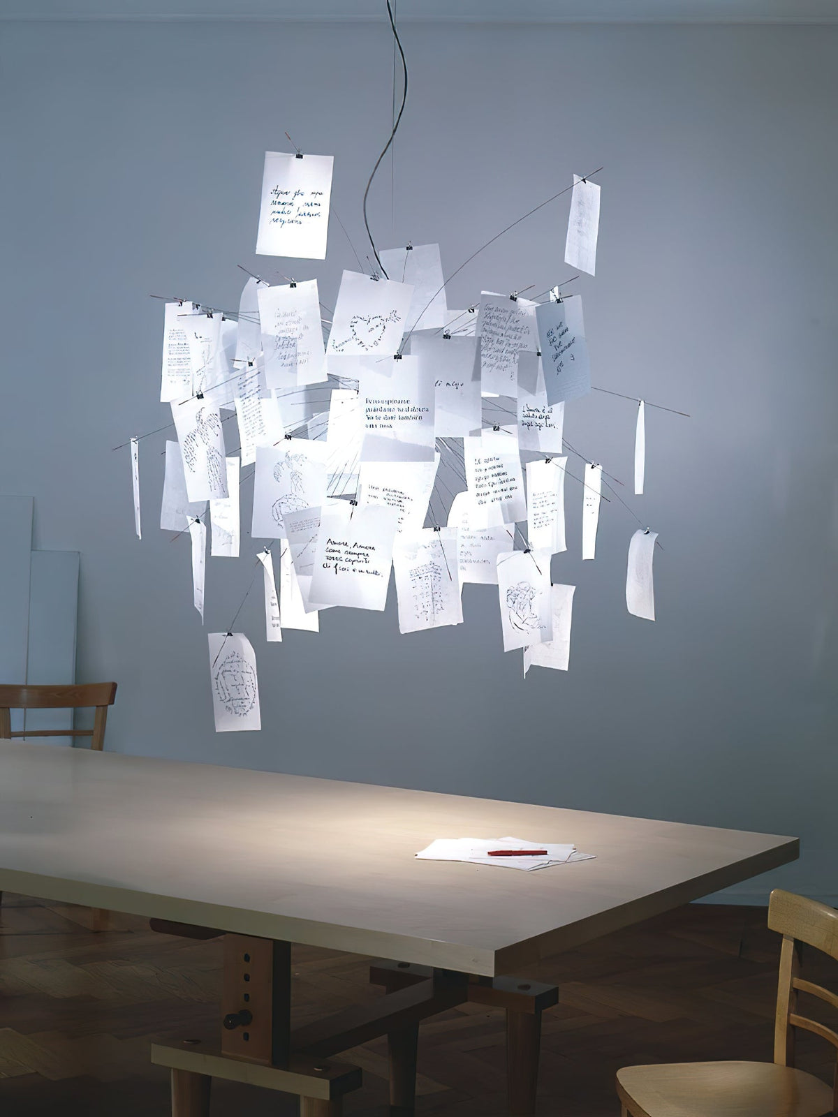 A Clérachic paper pendant over a table in a blue-grey room, framed on a white background, its notes softly lit.