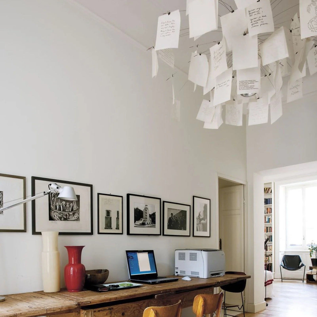 Above a wooden desk with a lamp and a framed print gallery, this Clérachic paper pendant spills notes across the ceiling.