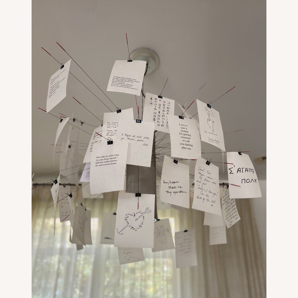 From below, a Clérachic paper pendant with handwritten cards clipped to red-tipped wire arms hangs against a window with sheer curtains.