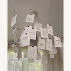 From below, a Clérachic paper pendant with handwritten cards clipped to red-tipped wire arms hangs against a window with sheer curtains.