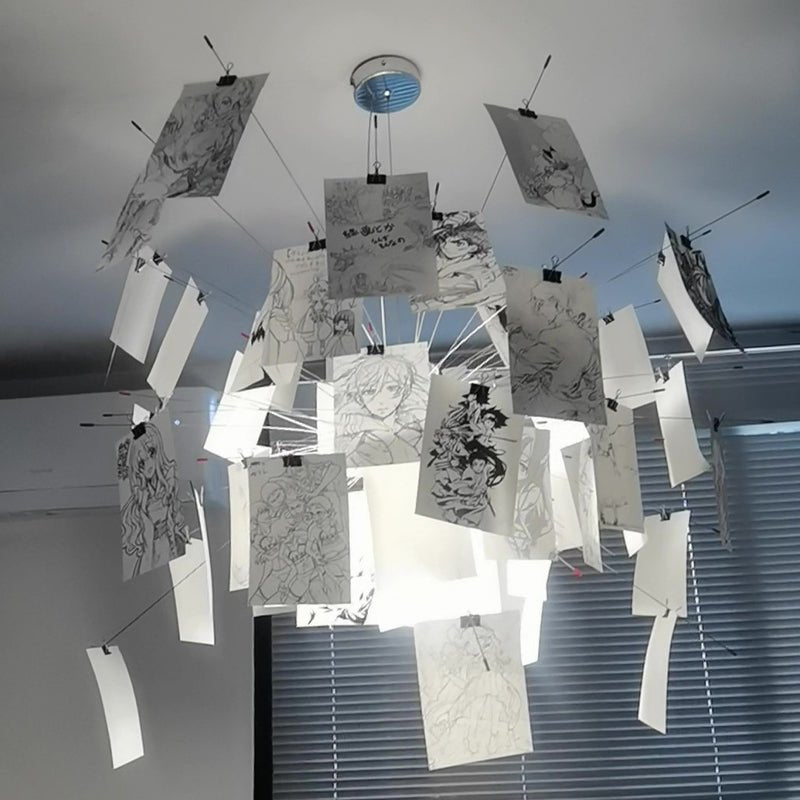 Black ink sketches of figures cover the cards on this Clérachic paper pendant, seen from below against blinds and a ceiling.