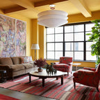 A Clérachic three-tier paper pendant hangs above a coffee table in a mustard-yellow room with large black-framed windows and a patterned rug.