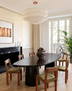 A Clérachic tiered paper pendant hangs in a calm dining room above a dark oval table with wooden chairs, beside tall French windows.
