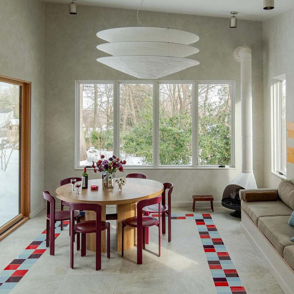 A Clérachic tiered white pendant hangs in a tall pale room with large windows, above a round table and red chairs on a tiled floor.