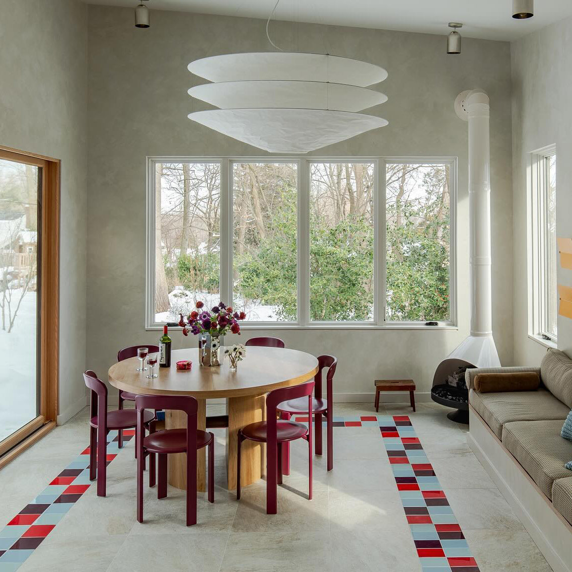 A Clérachic tiered white pendant hangs in a tall pale room with large windows, above a round table and red chairs on a tiled floor.