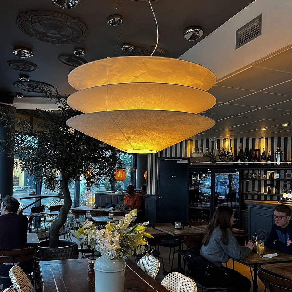 Glowing a warm amber, a large Clérachic tiered paper pendant hangs in a restaurant with an olive tree and tables behind.