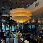 Glowing a warm amber, a large Clérachic tiered paper pendant hangs in a restaurant with an olive tree and tables behind.