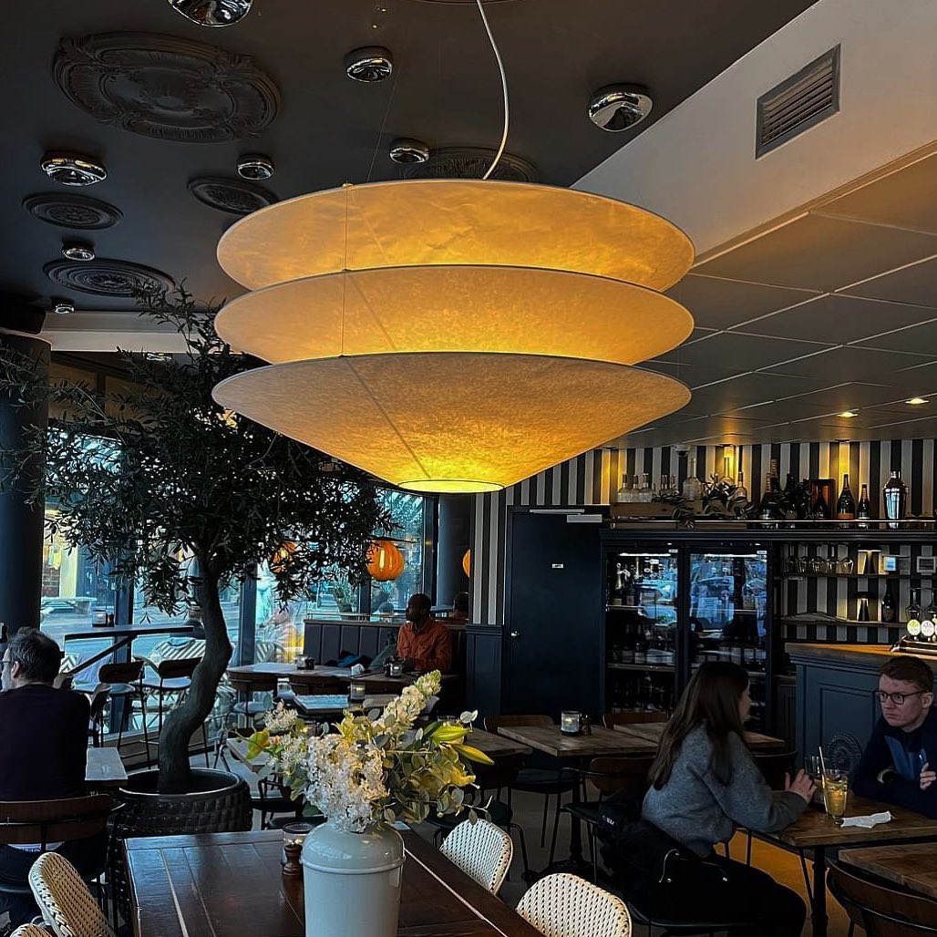 Glowing a warm amber, a large Clérachic tiered paper pendant hangs in a restaurant with an olive tree and tables behind.