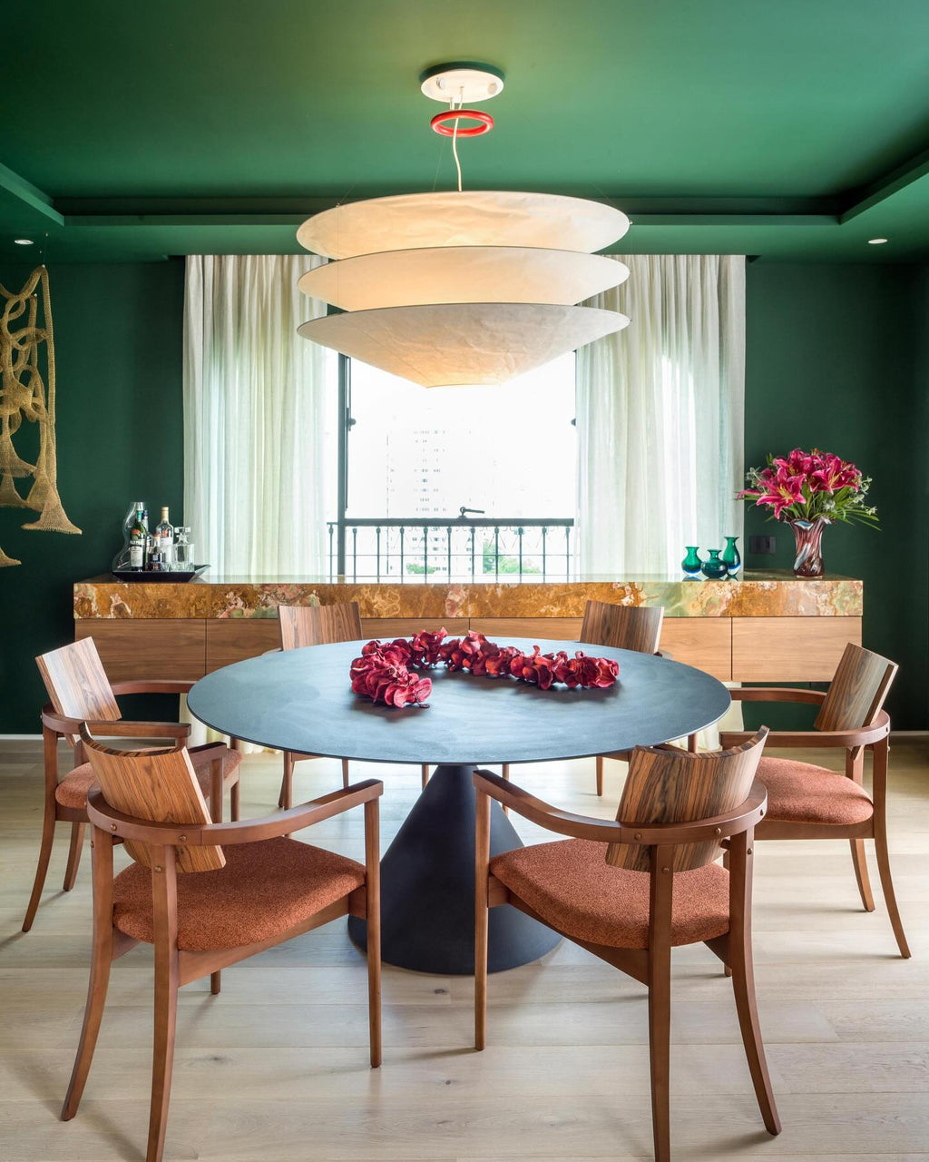 A Clérachic three-tier paper pendant hangs in a green-walled dining room above a round dark table with orange-cushioned wooden chairs.