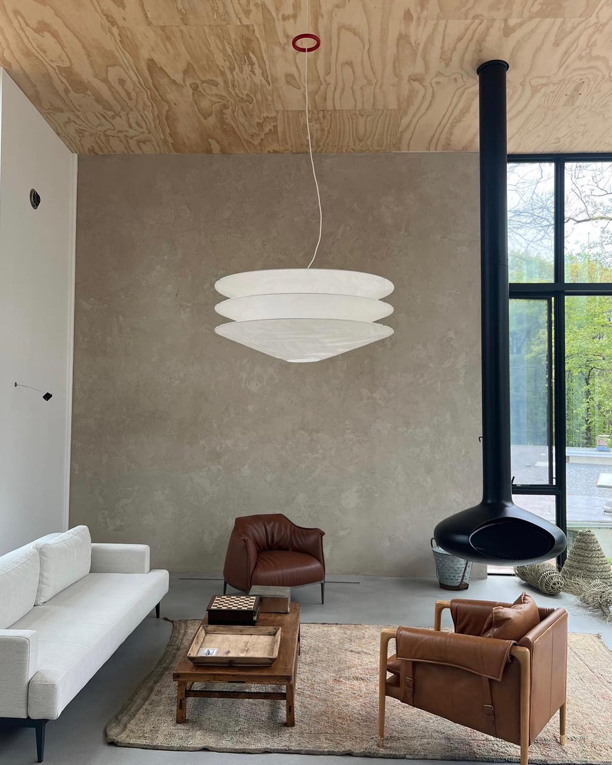 A Clérachic tiered white pendant hangs from a plywood ceiling in a plaster-walled lounge with a pale sofa, leather chairs and a hanging fireplace.