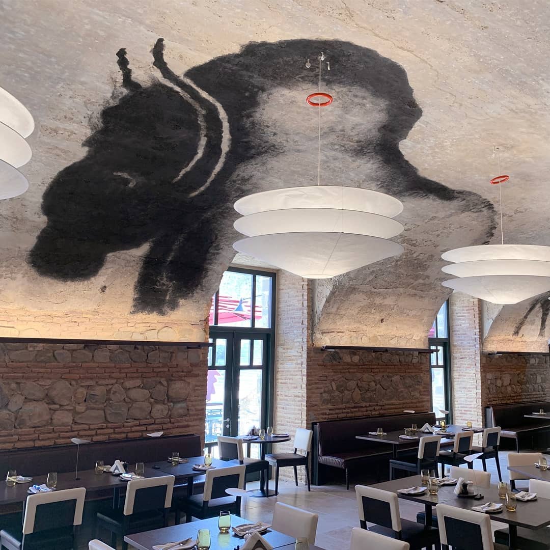 A Clérachic tiered paper pendant hangs in a vaulted brick restaurant with a large painted figure on the ceiling above the tables.